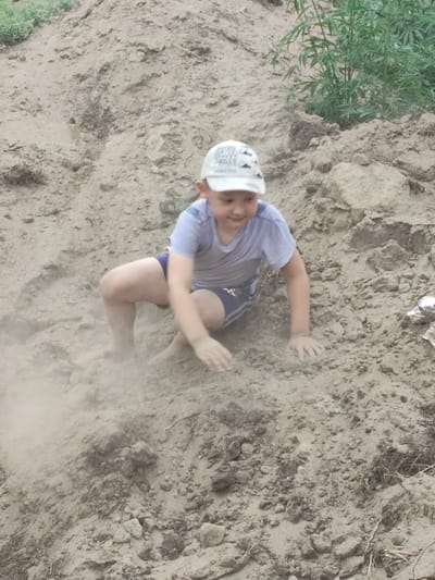 A young boy wearing a white baseball cap and a light blue t-shirt with dark blue shorts is playing in a pile of dirt. He is in a crouched position, with his right hand on the ground, raising dust. His left leg is bent, and his right leg is extended behind him. The ground is covered in dirt and rocks. In the background, there is some green vegetation. The scene appears to be outdoors, possibly in a park or a construction site.