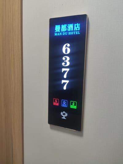 A vertical, black digital display with a gold border is mounted on a textured, light-colored wall next to a light-colored wooden door frame on the left. The display shows the hotel name "曼都酒店 MAN DU HOTEL" in blue text at the top. Below the hotel name, large white numbers "6377" are displayed vertically. At the bottom of the display, there are four icons. From left to right, the icons are: a red bell icon with Chinese characters and "DO NOT DISTURB" in English, a blue house icon with Chinese characters and "CHECK IN" in English, a green figure icon with Chinese characters and "PLEASE CLEAN UP" in English, and a black bell icon with Chinese characters and "DOORBELL" in English. The overall scene is static and appears to be an interior shot of a hotel hallway.
