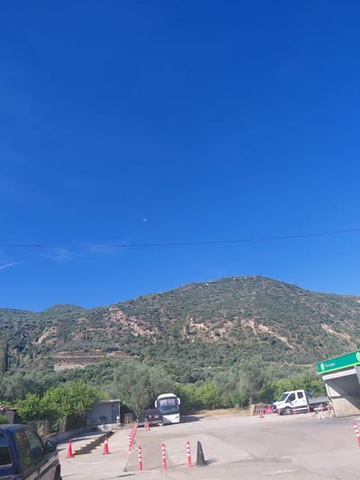 This image captures a wide outdoor scene in the Municipal Unit of Diakopto, Greece, under a clear, bright blue sky. A distinct crescent moon is visible in the upper center-right portion of the sky, with a faint contrail present in the upper-left. A thin, dark horizontal wire traverses the middle of the sky.

In the background, a large, prominent mountain range rises, densely covered in green scrub, trees, and some exposed rock formations. Terraced cultivation is visible on some of the slopes.

The midground and foreground depict a paved service or parking area at the base of the mountains. On the far left, the rear and side of a dark-colored pickup truck or SUV are partially visible. Towards the center-left, a small, grey building with a pitched roof is nestled among lush green trees, including some with orange fruits.

In the center, a white tour bus and a black SUV are parked facing right, while a white flatbed truck is parked facing left further to the right. A vehicle service area, possibly a car wash, is delineated by orange traffic cones with white stripes and red-and-white barrier chains. This area features a recessed section with yellow markings on the ground. To the right, a prominent green and white canopy structure, labeled "Πλυντήριο" (Car Wash) and featuring a 'Revoil' logo, covers what appears to be a car wash bay. Several wooden pallets are stacked near the white flatbed truck. No people are visible in the scene. The overall environment suggests a sunny, semi-rural setting.