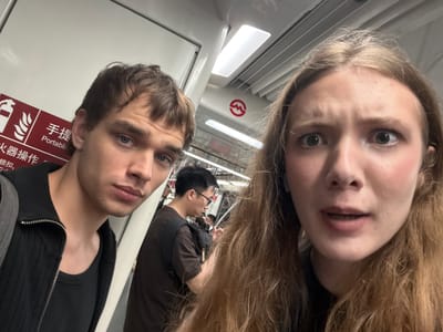 A close-up, selfie-style photograph taken on a public transit vehicle, likely a subway, within the Changning District of China. Two individuals, a man and a woman, are the primary subjects. The man, on the left, has short brown hair and blue eyes, and is wearing a black zip-up jacket over a black t-shirt. The woman, on the right, has long blonde hair and wide, surprised eyes, with visible blush on her cheeks. Her mouth is slightly open. In the background, another man with short black hair and glasses is visible, along with other passengers and the interior of the subway car, which features fluorescent lighting and a red metro logo on the wall. A fire extinguisher sign with Chinese characters is also visible to the left of the man.