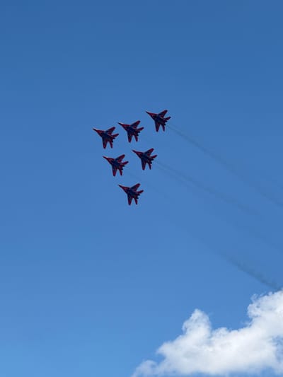 Six red and blue fighter jets fly in formation against a clear blue sky. The jets are arranged in a diamond shape with two jets leading, three jets in the middle row, and one jet trailing. They are leaving contrails behind them. The lower right corner of the image shows a few white clouds.