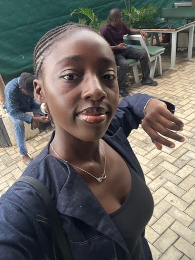 The image shows a selfie of a young Black woman with braided hair, wearing a dark blue denim jacket over a black tank top, a silver necklace with an infinity symbol pendant, and large silver hoop earrings. She has on braces on her teeth. In the background, another Black man is seated at a table with a maroon shirt and black pants, holding a mobile phone. To the left of the woman, another Black man wearing a blue checkered shirt and blue jeans is partially visible, bending over. The scene appears to be outdoors, with a paved walkway in the foreground and greenery in the background, suggesting a public space like a cafe or restaurant. The overall lighting suggests it is daytime. This setting could be consistent with outdoor seating areas found in various urban locations, including Karum, Nigeria, where such establishments often feature outdoor patios with paved surfaces and landscaping.