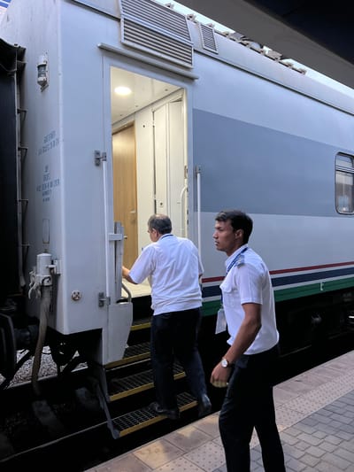 Two men, likely train conductors or railway workers, are standing on a train platform next to a train. Both men are wearing white short-sleeved collared shirts and dark pants. The man in the foreground, facing away from the camera and looking down towards the train car, is wearing a watch on his left wrist. He appears to be in the process of attending to something on the train. The man behind him, also looking down, is partially visible. The train appears to be a passenger train, with a grey exterior and blue, white, and red stripes along its side. The platform has a textured surface and yellow markings along the edge. The lighting suggests it is daytime, possibly late afternoon or early evening. The scene is set at a train station, with the environment suggesting an urban setting, consistent with Samarkand City, Uzbekistan. Visible text on the side of the train car appears to be in Cyrillic script, possibly indicating manufacturing or operational details. One visible piece of text reads "AO 781875 / 11.09.2020".