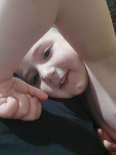 The image displays a close-up, low-angle shot of a young child lying on a dark surface, likely a bed or sofa. The child's head is tilted back, and their face is partially obscured by their arms. One arm is raised over their forehead, while the other is bent at the elbow and rests near their cheek. The child has fair skin, dark eyes, and is smiling with their mouth open, revealing teeth. Their lips appear to be lightly colored, possibly with lipstick. The background consists of a dark, textured surface that blends with shadows, suggesting an indoor setting. The lighting is soft, creating a warm glow on the child's face and arms. There is no identifiable information that would place this scene in Carbuna, Moldova; the description is limited to the visual elements present in the image.