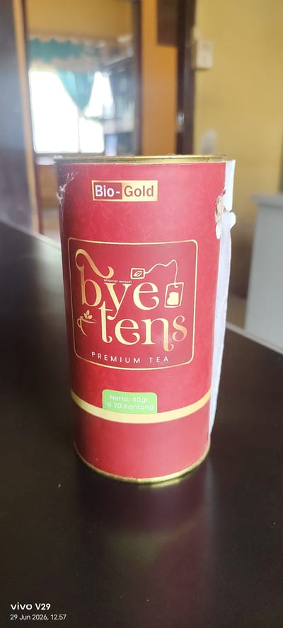 A close-up, eye-level shot shows a cylindrical red container of "bye tens" premium tea. The container has a golden lid and features the brand name in large golden letters, with a smaller line reading "Minuman rempah" above it. A gold label with "Bio-Gold" is at the top, and a green label at the bottom states "Netto: 40gr Isi 20 Kantong." The container is placed on a dark, reflective surface, possibly a table, in what appears to be an indoor setting with soft lighting. The background is blurred, showing what looks like a doorway or window with light filtering through. The timestamp in the bottom left corner indicates "vivo V29, 29 Jun 2026, 12:57". There are no people or animals in the image. The scene is devoid of any specific location markers within Pulodolang, Indonesia, or any other city.