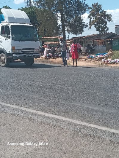 The image, rotated 90 degrees clockwise, captures a bustling roadside scene in Kipkarren, Kenya, under a bright, clear sky. A paved, two-lane road with a visible white dividing line runs diagonally across the frame.

Along what would be the left side of an upright image (appearing as the top side in the rotated view), a lively stretch features several informal shops and trees. A woman in a red dress stands near the road, accompanied by a man in a plaid shirt, both facing towards the cluster of shops. Other individuals are visible among the shops, some appearing to be seated or engaged in daily activities.

Prominently displayed on the shop fronts are signs for "M-PESA," a mobile money service, and a partially visible "SAFARIKOM SHOP," indicating a telecommunications provider. Various goods, likely textiles or other merchandise, are openly arranged outside these establishments.

Further along the roadside (in the lower-left section of the rotated image), a white truck, possibly an Isuzu model, is parked. Its license plate, "KBM 510Y," is clearly visible, and its hood appears slightly ajar or open. Several tall trees dot the landscape, offering partial shade to the area. The ground adjacent to the road is unpaved and dusty. The bright sunlight indicates it is daytime with good weather. A "Samsung Galaxy A04e" watermark is present in the bottom right corner of the image.