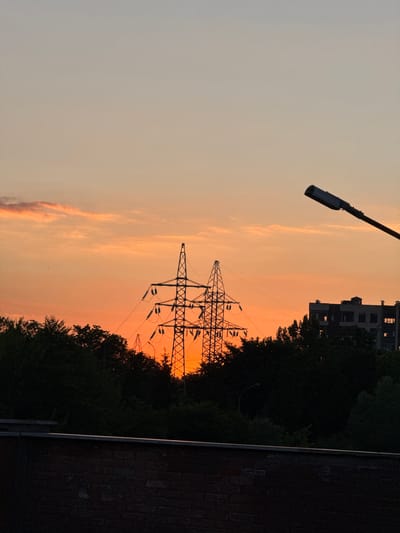 This media file shows a silhouette of power lines against a sunset sky. The sky is a warm gradient of orange and pink, with some wispy clouds. In the background, there is a building and a dense line of trees. A lamppost is visible in the upper left corner of the image. The overall mood is serene and peaceful, suggesting the end of a day. The time of day is clearly sunset, indicated by the colors of the sky. The location cues are limited, but the presence of power lines and apartment buildings suggests an urban or suburban setting. There is no visible text in the image.