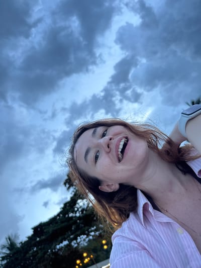 A close-up, upside-down selfie shows a woman with long, reddish-brown hair and freckles smiling broadly, revealing her teeth. She is wearing a pink and white striped button-down shirt and a gold necklace. A smartwatch is visible on her left wrist. The background is a mix of dramatic, cloudy skies and a blurred, dark green tree line with bokeh lights, suggesting an outdoor setting at dusk or during a cloudy day. The weather appears overcast. The overall emotion conveyed is cheerful and uninhibited. The location is identified as Baan Chaweng Noi, Thailand.