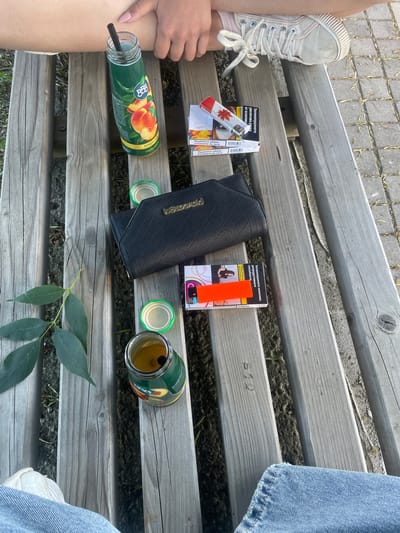 A person sits on a wooden park bench in Elin Pelin, Bulgaria. The person is wearing a light-colored sneaker and blue jeans. On the bench, there is a bottle of peach juice with a straw, an open cigarette pack, a lighter, and a black wallet. There are also two green bottle caps and a branch with green leaves. The bench appears to be outdoors, with a stone or paved surface visible beyond the bench.