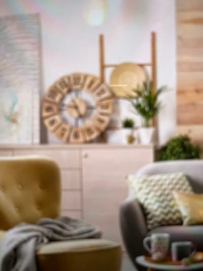 The image displays a room's interior, featuring a wooden clock on a wall with shelves holding a plate and greenery. A grey chair with a patterned pillow and a side table with a mug and a device are positioned near the wall. A mustard-colored chair is partially visible, draped with a grey blanket. The image is blurred.