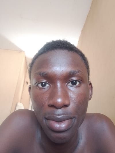 A close-up, self-portrait of a young Black male with dark skin, short, dark hair, and dark eyes. The background appears to be an interior room with beige walls and a white ceiling. The subject is the only person visible in the image. Specific location within Panhauya, Nigeria cannot be determined from the image.