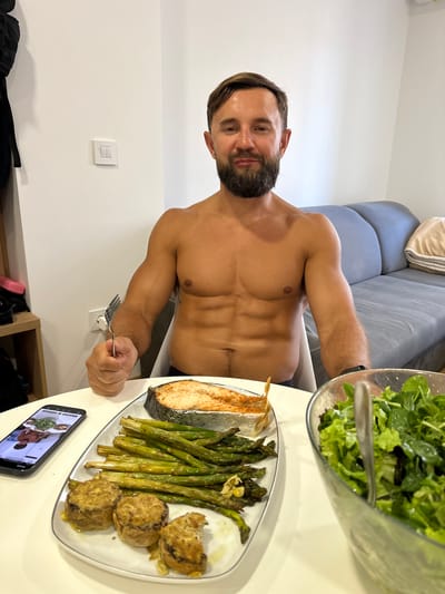 A shirtless man with a beard is sitting at a table in Donji Pobori, Montenegro, in front of a meal. The meal consists of a piece of salmon, roasted asparagus, what appear to be stuffed mushrooms or patties, and a bowl of salad. A smartphone is also on the table, displaying an image of a man holding a salad. The man in the image is holding a fork in his right hand, seemingly about to eat. In the background, a grey sofa and a white wall are visible.