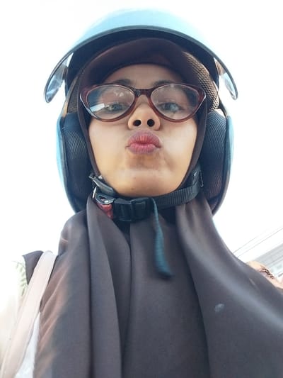 A low-angle, close-up selfie shows a young woman wearing a blue motorcycle helmet and a dark brown hijab. She has on cat-eye glasses with brown frames and is puckering her lips, which are painted with red lipstick. The background is a bright, overcast sky. Her shoulders and chest are covered by the hijab, and a light-colored strap of a bag is visible on her left side. The image is taken outdoors, possibly in a public space in the city of Macege, Indonesia.