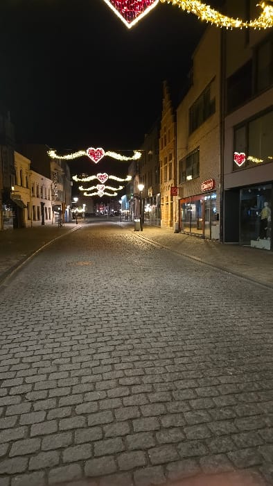 The image depicts a cobblestone-paved street at night in Herentals, Belgium. The street extends into the distance, flanked by multi-story buildings on both sides. Overhead, numerous strings of yellow-gold festive lights are draped across the thoroughfare, interspersed with large, illuminated red heart-shaped decorations. One prominent heart is positioned near the top of the frame. Additional streetlights with globe fixtures illuminate the cobblestone surface.

On the right side of the street, buildings feature commercial storefronts, some with large display windows. One shop window exhibits at least two mannequins dressed in clothing. A building on the right also displays a smaller illuminated red heart decoration on its facade and has a distinctive stepped gable roofline. Buildings on the left appear to be older structures, possibly residential or mixed-use, with multiple windows and architectural details. The street is devoid of people or vehicles, presenting a quiet scene under artificial illumination. No discernible actions or interactions are taking place.