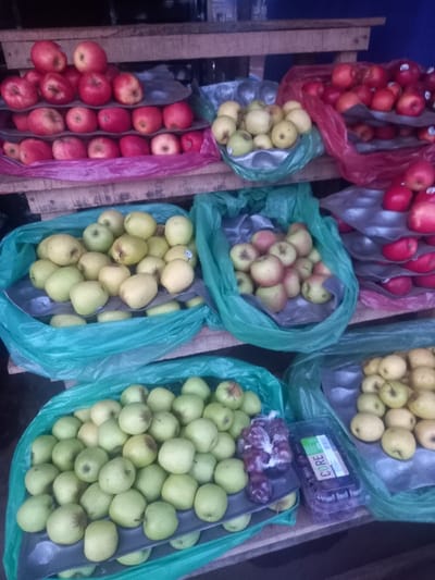 The image shows a fruit stand located in Akure, Nigeria. The stand is made of wooden planks with shelves arranged vertically. The shelves are filled with various types of fruits, including red apples, green apples, and yellow pears. Some of the fruits are contained in clear plastic bags. The fruit stand is located outdoors, and the background consists of a brick wall.