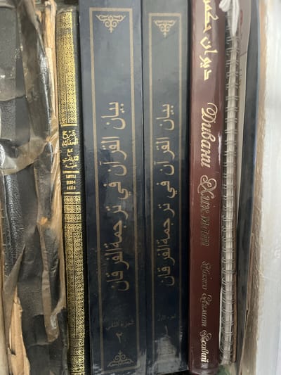The image shows a close-up of several books stacked vertically. The books appear to be old and worn, with visible signs of age and use. The spines of the books are decorated with intricate gold patterns. The titles are in Arabic script, suggesting they are religious texts. One book has a maroon cover with gold lettering that appears to be in Latin script, possibly a translation or commentary on the Arabic texts. A spiral-bound notebook is also visible, partially obscured by the books. The lighting is dim, and the background is out of focus, making it difficult to determine the exact environment. However, the presence of stacked books suggests a library, study, or personal collection. There are no people or animals in the image, and no specific location details can be inferred beyond the general context of books being stored. The overall impression is one of scholarly or religious material.