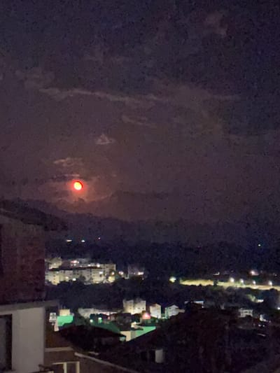 The image presents a dark, blurry night scene, oriented vertically as if rotated clockwise. The overall impression is of a low-light environment with considerable visual noise.
A prominent, circular, intensely red light source dominates the left-central portion of the frame. It appears to be glowing brightly against a dark, textured background that resembles a cloudy or hazy night sky, with the light partially obscured by these atmospheric conditions. This red object is likely the moon, appearing red due to atmospheric particles.
The right side of the image depicts a city or town at night, viewed from an elevated vantage point. Numerous bright, artificial lights illuminate the urban landscape. A distinct, long, vertical streak of lights suggests a road or thoroughfare, while scattered clusters of lights indicate buildings and other city structures. Faint outlines of dark buildings or rooftops are visible along the far right edge and bottom, suggesting the viewpoint is from a building or balcony overlooking the area.
No individuals or discernible text are visible. The time of day is clearly night, underscored by the pervasive darkness and the reliance on artificial light sources. The atmospheric conditions appear hazy or cloudy, contributing to the image's overall softness and the reddish appearance of the circular light. The scene captures a nighttime view over Sandanski, Bulgaria.