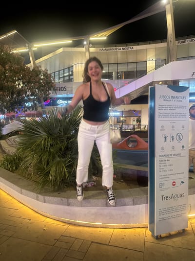 This is a candid, nighttime photograph taken outdoors in what appears to be a modern shopping or entertainment complex. A young woman is captured mid-stride, her body angled towards the camera with a wide smile. She is wearing a black tank top and white pants, with black and white Converse sneakers.
Behind her, signs for "HAMBURGUESA NOSTRA" and "PURE CUIS" suggest food establishments. Another prominent sign is for "JUEGOS INFANTILES" (Children's Games) and "TresAguas," a water park, with operating hours listed for both mornings and afternoons during summer. The sign also includes rules and contact information, such as emergency services. The presence of "VIPS" and "GINOS" logos indicates other businesses within the complex.
The lighting suggests it is evening, with artificial lights illuminating the walkways and building facades. A pathway is visible, constructed with stone tiles, and bordered by greenery, including palm fronds and flowering bushes. The overall atmosphere is lively, likely a recreational area open in the evening. The woman's joyful expression and dynamic pose suggest she is enjoying herself in this vibrant setting.