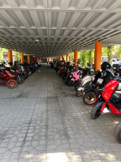 The image depicts a parking area in Petulu, Indonesia, densely packed with scooters of various colors and models. The parking area is covered by a gray corrugated metal structure supported by orange beams. A person wearing a white shirt and dark shorts is visible walking down the center of the parking area. The ground is paved with gray stone tiles. The scooters are lined up in rows, filling the parking space. Foliage is partially visible between the support beams of the parking structure.