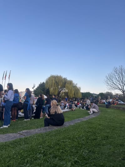 The image depicts an outdoor gathering in Mirandela, Portugal, occurring during late afternoon or early evening under a clear, light blue sky.
The scene features a public green space with a large congregation of individuals. In the immediate foreground, vibrant green grass is visible, traversed by a curved, multi-tiered stone retaining wall or path. A person with blonde hair, wearing dark clothing, sits on one of these low stone walls with their back to the viewer, engaged with a mobile phone. To the left, a woman in a light blue shirt and dark trousers stands near a camera mounted on a tripod, facing towards the right.
In the midground, a significant crowd, predominantly young adults, is distributed across the grassy area and seated on stone structures. Many individuals are observing towards the right side of the frame, suggesting a focal point outside the immediate view. Some are conversing while others are using mobile phones. A large weeping willow tree with dense, light green foliage dominates the left-midground, while a bare deciduous tree with fine branches stands on the right.
The background reveals a series of buildings, including one with a "RESTAURANT" sign, partially obscured by the crowd and foliage. Three flagpoles are visible on the far left, displaying flags with vertical green, red, and yellow stripes, consistent with Mirandela's municipal colors. Vehicle traffic or parked vehicles are minimally visible behind the bare tree on the right. No discernible specific events or interactions beyond general gathering are evident.