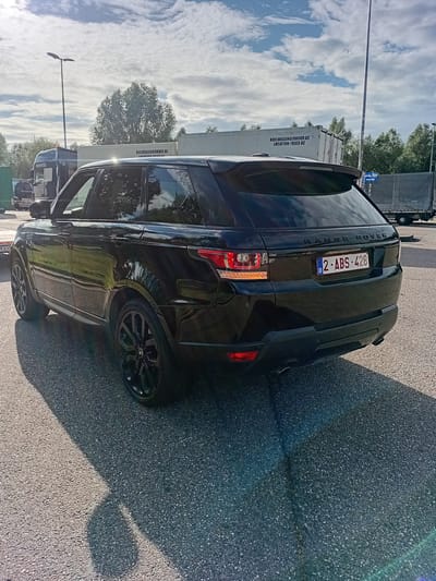 This is an eye-level, slightly tilted, outdoor photograph of a black Range Rover parked on an asphalt surface. The vehicle is the primary subject, viewed from its rear passenger side. The car has a license plate that reads "2 ABS 428 B". The background consists of a light-colored trailer or container with text that appears to say "KOELWAGENVERHUUR.BE LOCATION-TRIGOBE". Beyond the trailer, trees and a bright blue sky with fluffy white clouds are visible. A street light pole is also present in the scene. The sunlight creates distinct shadows on the asphalt, indicating it is daytime, likely afternoon, and the weather is sunny. The scene appears to be a parking lot or roadside area, possibly in an industrial or commercial zone, given the presence of the trailer and trucks in the distance. No people are visible in the frame.