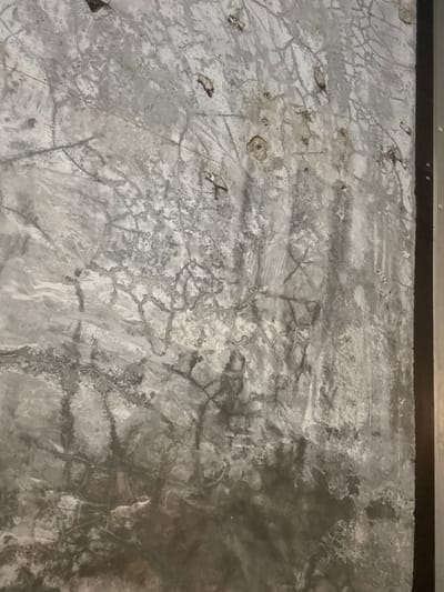 This image depicts a detailed view of a highly textured and weathered wall, located in Ko Tao, Thailand. The wall surface is predominantly gray, exhibiting a varied palette from light ash-gray to dark charcoal, with a mottled and aged appearance. A dense, irregular network of fine, dark cracks, resembling a spiderweb pattern, is visible across the entire surface. Numerous small, uneven indentations, chips, and areas of exposed subsurface material are scattered throughout, with several metallic, circular fasteners or remnants of such objects clustered in the upper-right and central regions. Dark, vertical streaks and blotches of discoloration are prominent, particularly in the lower and central sections. A distinct dark, vertical planar element, possibly a frame or architectural trim, with a thin, lighter-colored inner edge, is visible along the right side of the image. No individuals, animated subjects, or active interactions are present in the scene.