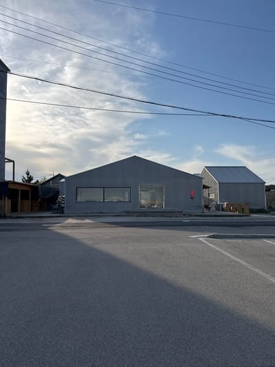 The image depicts a street scene in Toseong-myeon, South Korea. Two corrugated metal buildings are visible. The first, a gabled structure, features a large rectangular window and a smaller window above. A red neon sign is affixed to the building's facade. The second building is partially visible behind the first. A paved road stretches alongside the buildings, exhibiting lane markings and a shadow cast upon it. Overhead, power lines cut across a sky marked by scattered clouds. A rudimentary wooden structure is also visible in the foreground.