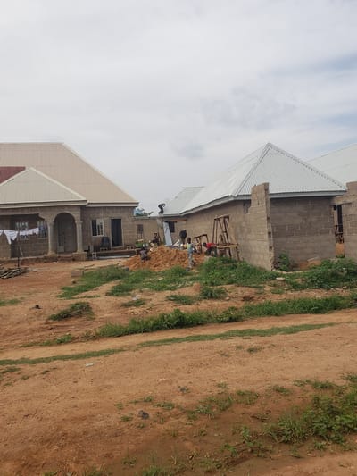 The image depicts an outdoor scene in the city of Kachia, Nigeria, featuring residential construction. In the foreground, a dirt path with scattered green grass and some debris leads towards two newly constructed houses. The house on the left is more complete, with a finished, light-colored corrugated metal roof, visible columns and archways at the entrance, and windows. Laundry is hanging on a line in front of this house. The house on the right is still under construction, with walls made of grey cinder blocks and a similar corrugated metal roof in place. Several people are engaged in construction work. One person is working on the roof of the house on the right. Others are gathered around a pile of sand near the house on the right, possibly involved in mixing or transporting materials. The environment is characterized by a cloudy sky, dry earth, and sparse vegetation.