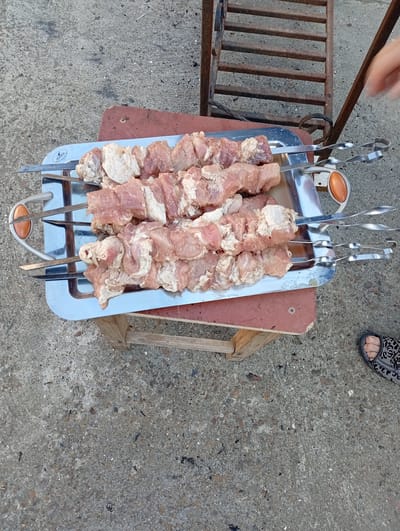 In Buțeni, Moldova, on a concrete surface, a metal tray holds several skewers of raw, cubed meat. The meat appears to be pork, with visible fat and marbling. The skewers are made of metal and are arranged horizontally across the tray. The tray itself is rectangular with rounded corners and silver handles. It rests on a wooden support, which is placed on the concrete ground. In the background, a rusted metal grill structure is visible. A hand with blurred motion is seen reaching towards the grill. A person's foot, wearing a patterned sandal, is also visible on the lower right side of the frame. The overall scene suggests preparation for outdoor cooking.