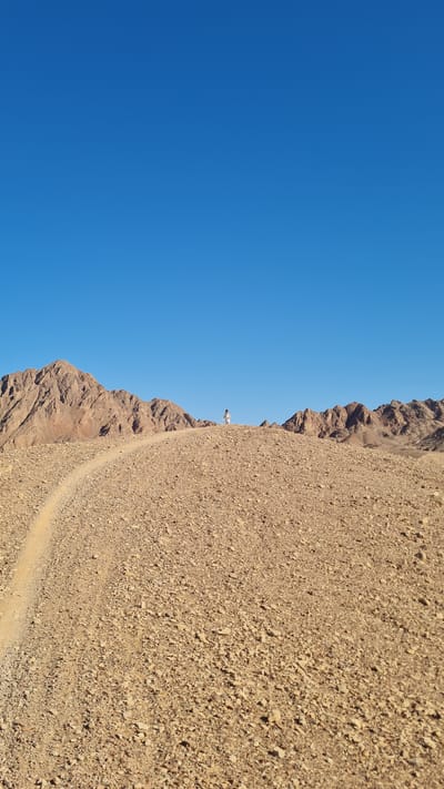 The image depicts a desert landscape in Eilat, Israel. A steep, rocky mountain slope dominates the right side of the frame, composed of loose brown and beige gravel and rock. A narrow, winding dirt path is visible on the mountainside.
To the left of the mountain slope is a clear blue sky. A single person, dressed in white, stands near the edge of the mountain.