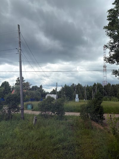 This media file shows a rural outdoor scene in Гаревая, Russia. The image is oriented vertically. In the foreground, a wooden utility pole with wires is prominent. Beyond it, a dirt path runs alongside a grassy area and dense trees. In the middle ground, a small electrical transformer box is visible, along with more trees and a larger metal communication tower in the distance. The sky is overcast and appears dark with heavy clouds, suggesting a stormy or gloomy weather condition. The time of day is indeterminate due to the cloud cover, but it appears to be daytime. There is no visible activity or people in the frame. There is some text on a green sign near the path in the middle ground, but it is not legible. The overall scene conveys a sense of remoteness and undeveloped infrastructure, typical of a rural setting.