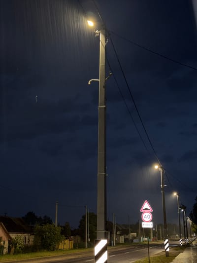 The image captures a nighttime scene during heavy rainfall in Лабжанскі сельскі Савет, Belarus. The sky is dark blue and overcast, with visible streaks of rain illuminated by the artificial light sources.
A paved, two-lane road runs through the center and foreground of the image, appearing wet from the rain. White road markings and yellow rectangular reflectors are visible on its surface. To the right of the road, a wet, paved sidewalk is present, lined with reflective white and black striped bollards.
Multiple utility poles, likely made of concrete or metal, stand along both sides of the road, supporting an array of overhead power lines and cables. Each pole features streetlights that emit a warm, yellowish glow, brightly illuminating the falling rain directly beneath them.
Prominently featured on the right side of the road is a set of reflective traffic signs. The topmost sign is a red-bordered triangle indicating a speed bump. Below it, a red-bordered circular sign shows "40," denoting a 40 km/h speed limit. The lowest sign is a white rectangle with an upward arrow and "↑100M↑," indicating the speed bump is 100 meters ahead. A large white and black striped reflective chevron marker is affixed to the leftmost utility pole.
On the left side of the road, several older-style residential houses are visible, partially obscured by trees and bushes. One house has a reddish-brown roof and light-colored walls, while another shows a yellow fence. The surrounding vegetation, including trees, bushes, and grass, appears dark and somewhat blurred due to the low light and rain.
In the upper left portion of the frame, near the primary streetlight, faint, squiggly light trails are present, suggesting motion blur of small objects or particles caught in the light. No human figures are visible in the scene.