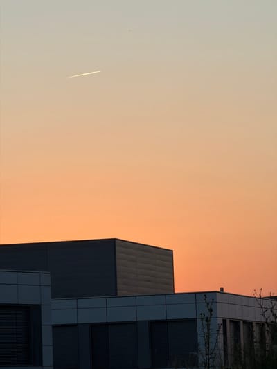 An aerial view of a modern office building in Leudelange, Luxembourg, at sunset. The sky displays a gradient of warm orange and yellow hues. A faint contrail from an aircraft is visible in the upper left portion of the sky. The building features a multi-level design with dark gray and white facades and prominent horizontal windows, many of which are covered by closed blinds. Several barren plants are visible in the foreground, silhouetted against the warm sky.