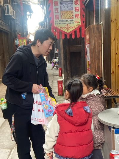 An outdoor scene depicts a man in a black jacket holding toys in a white bag. Two young girls are nearby, one in a floral dress and the other in a red vest. A banner advertises local food items, including "Ban Ci Ba" (rice cake) and smoked tofu. The streetscape features traditional Chinese architecture, including wooden buildings and red mailboxes. Text on the banner shows a phone number and address in Chongqing.