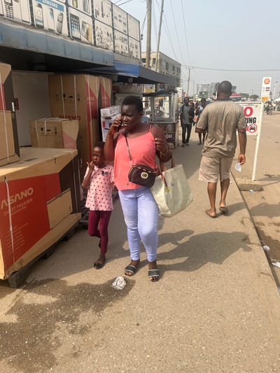 The image portrays a daytime urban street scene in Ablekuma, Ghana. In the foreground, an adult female with dark complexion, wearing a coral sleeveless top, light blue jeans, and black and white slide sandals, is engaged in a phone conversation, holding a black smartphone to her right ear. She carries a black crossbody bag with a gold emblem and a large light-colored tote bag. Standing to her left is a young dark-complexioned child, dressed in a pink polka-dotted short-sleeved top, dark red leggings, and black flat shoes; her right hand is near her mouth.

To the immediate left of these individuals are stacked cardboard boxes, some explicitly labeled "ASANO CHEST FREEZER Model: AS-500," indicating retail appliances. Behind these boxes, a storefront displays multiple rectangular advertisements for mobile phones and tablets, surmounted by a blue awning.

In the mid-ground, an adult male with dark complexion walks away from the camera, wearing a light brown short-sleeved shirt, matching shorts, and dark flip-flops. He holds a small white object in his right hand. Adjacent to him, a white circular sign with a red border and a car symbol indicates a "NO PARKING" zone, next to a vertical pole displaying a red "SHELL" logo.

The concrete sidewalk exhibits wet patches and scattered litter, including a crumpled white object near the primary woman's feet. Distinct, long shadows are cast by the individuals on the ground, indicating direct sunlight. In the background, several additional pedestrians are visible, along with multiple multi-story buildings and an asphalt roadway under a clear, hazy sky.