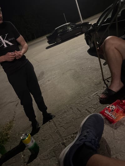 The image shows a group of people in what appears to be an outdoor setting at night. One person is wearing black sweatpants and a black t-shirt with "Calvin Klein Jeans" visible on it. Their hands are clasped in front of them, and they are wearing a ring. Another person is wearing black flip-flops and has their legs crossed. A package of "Cometų salty sticks" with a panda graphic is visible, along with a green and yellow can. A black car is parked in the background. The ground is paved with cobblestones or concrete. The scene suggests a casual gathering or break.