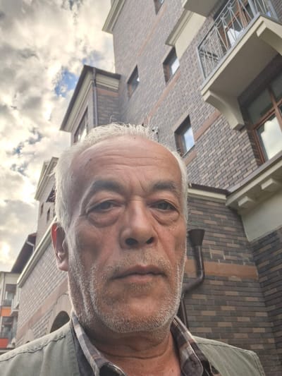 A man with graying hair and a stubbled beard takes a selfie from a low angle, looking towards the camera. He is outdoors, with buildings featuring brickwork and windows visible in the background, as well as a cloudy sky. The man appears to be wearing a collared shirt and a vest or jacket. The image is taken in Vidnoye, Russia. The time of day is likely daytime due to the bright sky. The overall mood is neutral. There is no visible text in the image.