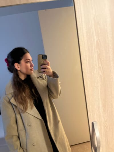 The image displays an indoor mirror selfie featuring a single adult female. The female, positioned on the left side of the frame, is seen in profile, looking towards the right. Her dark hair is partially pulled back, secured with a reddish-pink claw clip. She wears a light beige or grey double-breasted overcoat over a dark garment. A gold-toned ring is visible on her left ring finger. She holds a smartphone in her left hand, which has a light brown or tan case with a visible design, capturing her reflection in the mirror. The background reflected in the mirror consists of a segment of a light blue wall on the upper left and a lighter, off-white or cream-colored wall to the right. On the far right, a light-colored wooden surface, consistent with a door or wardrobe, is visible, featuring a gray rectangular handle. The setting is an interior space. The specific location within the city of Rennes, France, is not visually identifiable from the image.