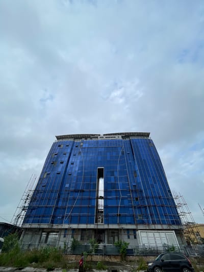 A low-angle, full shot captures a large, blue glass-paneled building under construction in Ilado, Nigeria, against a cloudy sky. The building has a distinctive, curved, trapezoidal shape, wider at the bottom and narrowing towards the top, with a central vertical cutout revealing scaffolding. Scaffolding structures are visible on both sides of the building, suggesting ongoing work. Multiple windows on the facade are open. At the very top, a flat roof structure with a crisscross pattern is present, potentially a helipad or rooftop amenity. In the foreground, a patch of overgrown green vegetation and some dirt paths are visible. A person in a pink shirt and dark pants walks on a path to the left of the frame, and a black SUV is parked on the right side. The overall environment appears to be an urban or semi-urban setting undergoing development.