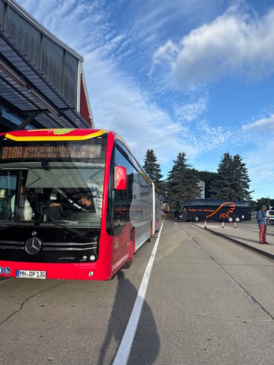 This image was taken at the bus station in Künersberg, Germany. In the foreground, a red Mercedes-Benz eCitaro bus is parked. The bus has a digital display showing "München Hbf Bahnhof". A license plate reading "MN DP 130" is visible on the front of the bus. The driver, a man wearing a dark shirt, is seated inside the bus, seemingly operating the vehicle. In the background, a black coach bus with orange accents is parked. This coach displays "Stuttgart - Ulm - Memmingen" and "ALLGÄU AIRPORT EXPRESS". A person in red pants and a blue jacket stands near the coach bus. The scene is set against a backdrop of a bright blue sky with scattered clouds and several tall evergreen trees. The environment appears to be an open-air bus terminal with asphalt ground and a building structure partially visible on the left.
