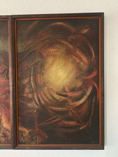 The scene depicts an abstract oil painting, which is part of a larger multi-panel artwork, mounted on a plain white wall in the city of Bogdanci, North Macedonia. The primary subject is the rightmost rectangular panel, framed in dark brown wood with a lighter inner trim. The artwork features a central luminous yellow-gold area, surrounded by swirling and textured layers of dark brown, deep red, and orange pigments. Raised, wavy lines, possibly applied with thick impasto or a cord-like material, crisscross the composition, with visible paint drips and splatters. A faint signature and the year '83 are present in the lower right corner of the right panel. A partial view of an adjacent, similarly framed panel is visible on the left edge. The wall behind the artwork is unadorned and white. No individuals, actions, or other objects are visible within the frame.