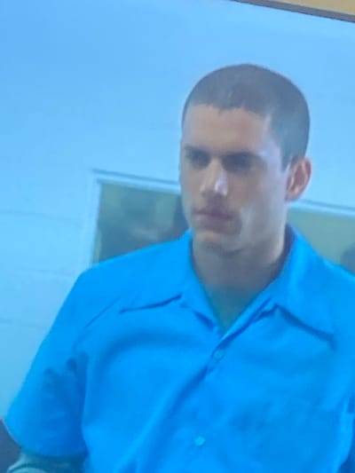 The image features a young man captured from the chest up, positioned against a light-colored, possibly indoor background that includes what appears to be a wall and a window or doorway. The man has very short, dark hair in a buzz-cut style and a medium complexion. He is wearing a light blue collared shirt. His expression is neutral, appearing somewhat serious or pensive, and he is looking slightly towards the viewer. The image is taken at a slight angle, with the man's head tilted. The lighting in the scene is gentle, casting subtle shadows on his face and clothing. There is no discernible specific activity taking place other than being photographed. The image quality is somewhat soft and grainy. No text is visible. While the location context is Gosa, Nigeria, no specific visual cues within the image confirm this.