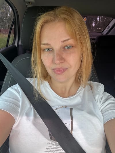 A close-up, slightly angled, medium shot captures a young Caucasian woman with blonde hair, fair skin with freckles, and green eyes, looking directly at the camera. She is seated in the driver's seat of a car, indicated by the presence of a black seatbelt crossing over her white t-shirt. The t-shirt has a printed design featuring text and an image of a building. The background is blurred, showing the interior of the car, including a black headrest and a partial view of the passenger side window with trees and foliage visible outside. The lighting suggests it is daytime. The image is an outdoor, daytime, candid photograph taken inside a vehicle. The specific location of Vereteya, Russia is not evident from the visual information provided in the image.