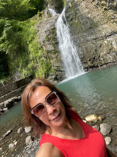 The image is a close-up, selfie-style shot of a woman in a red top and large, stylish sunglasses. She is positioned to the right of the frame, with her head tilted towards the camera, a slight smile on her face, and her mouth slightly open. Her brown hair is visible around her face and shoulders.
Behind her and to the left, a waterfall cascades down a rocky cliff face. The water is bright white and frothy as it falls into a calm, greenish-blue body of water. Lush green vegetation, including trees and grasses, grows on the left side of the waterfall and at its base. The foreground on the right side of the image shows a rocky shoreline with various sized stones of different colors, some submerged in the clear water. There are some dark, charred-looking stones or wood near the bottom right.
The lighting suggests it is daytime, likely sunny given the bright reflections on the water and the woman's sunglasses. The overall scene appears to be a natural setting, possibly a park or a nature reserve, given the waterfall and greenery. The location cues (Khostinskiy, Russia) combined with the natural beauty suggest a scenic outdoor environment. The woman's relaxed pose and smile convey a sense of enjoyment and contentment in the natural surroundings. No visible text is present in the image.