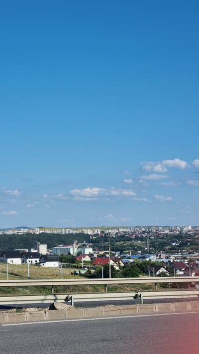 The image depicts a panoramic view of the city of Șcheia, Romania, under a clear blue sky with scattered white clouds. In the foreground, a multi-lane highway with a central barrier is visible, composed of asphalt and concrete. Beyond the highway, a verdant landscape unfolds, dotted with residential buildings. These structures range from modern single-family homes with dark roofs to multi-story apartment buildings forming the skyline of the city in the background. The residential area is nestled amongst rolling hills covered with trees. The overall scene suggests a developed urban area integrated with its natural surroundings. There are no people or animals visible in the frame.