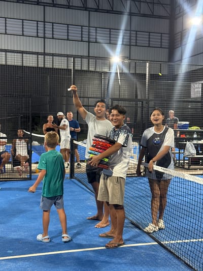 The image depicts an indoor padel court with a blue surface, enclosed by black wire fencing. Several individuals are present on the court, some appearing to be in a celebratory or awards ceremony. In the foreground, a young boy in a teal t-shirt and blue shorts stands facing away from the camera. To his right, a man in a grey t-shirt is holding a stack of padel rackets. Next to him, another man in a patterned t-shirt and khaki shorts smiles broadly. To the right of the court, a woman in a black and white t-shirt and shorts smiles and holds a padel racket. In the background, other individuals are visible, some watching the scene. The environment appears to be a sports facility with artificial lighting.