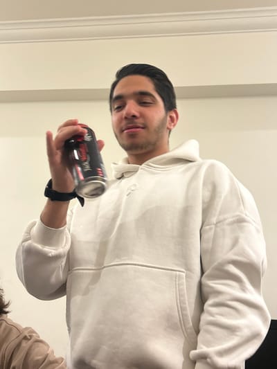 A young man in a white hoodie holds a black and red "Jack Daniel's" canned drink. He has dark hair and appears to be indoors. The background is a plain white wall and ceiling. Part of another person's shoulder and hair are visible in the lower right corner. He is also wearing a black wrist watch.