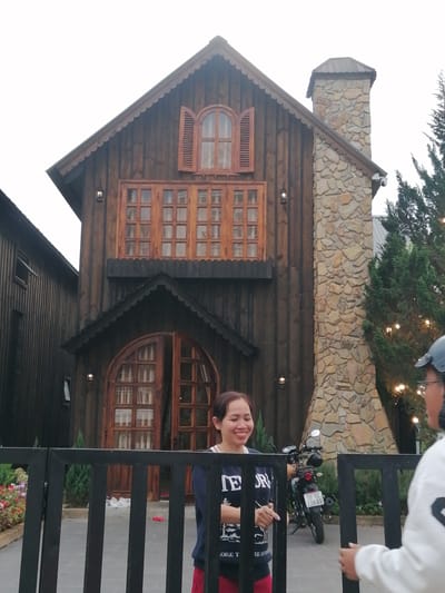 A woman with dark hair and a wide smile is standing behind a black metal fence in front of a rustic wooden building. She is wearing a dark blue long-sleeved shirt with white text and red pants. To her right, a motorcycle is parked. Behind her, the wooden building has multiple windows with brown frames and shutters, and a stone chimney. To the right of the building, evergreen trees are visible with decorative lights. The background is a bright, overcast sky. The location appears to be Lam Vien Square in Da Lat, Vietnam, a popular tourist spot known for its distinctive architecture and attractions.
