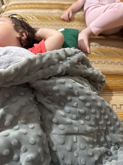 Two young children are asleep on a bed. The child on the left is wearing pink pajamas and is partially covered by a light-colored blanket with a pattern of raised dots. Their legs and feet are visible. The child on the right is mostly covered by a grey minky blanket with a textured dotted pattern. Only their head, with dark curly hair and an ear adorned with a small earring, is visible, along with a glimpse of their arm and torso clad in an orange shirt. Both children appear to be resting peacefully, their faces obscured or turned away. The background consists of a bedspread with a striped pattern in shades of yellow and brown. The lighting suggests it could be daytime, with soft shadows cast on the bedspread. The scene is intimate and domestic, depicting a moment of rest. There is no visible text in the image.