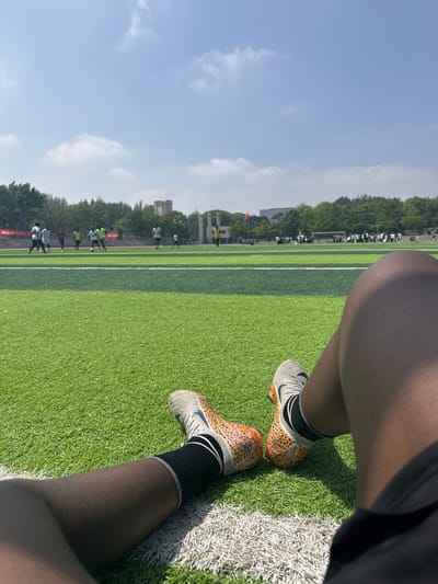 A ground-level, medium-angle shot shows a partial view of a soccer field on a sunny day. In the foreground are the legs and feet of a person wearing soccer cleats with a grey and orange cheetah print. The person is wearing black socks with white trim. The legs are dark-skinned and appear to be resting on the field. The soccer field is green with white boundary lines. In the background, other people are present, and some are actively engaged in what looks like a soccer training activity. Trees and buildings are visible in the distant background. The scene is in Linghe, China.